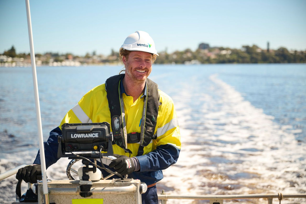 A man wearing a hard hat and safety vest stands on a boat, overseeing operations in a maritime environment.